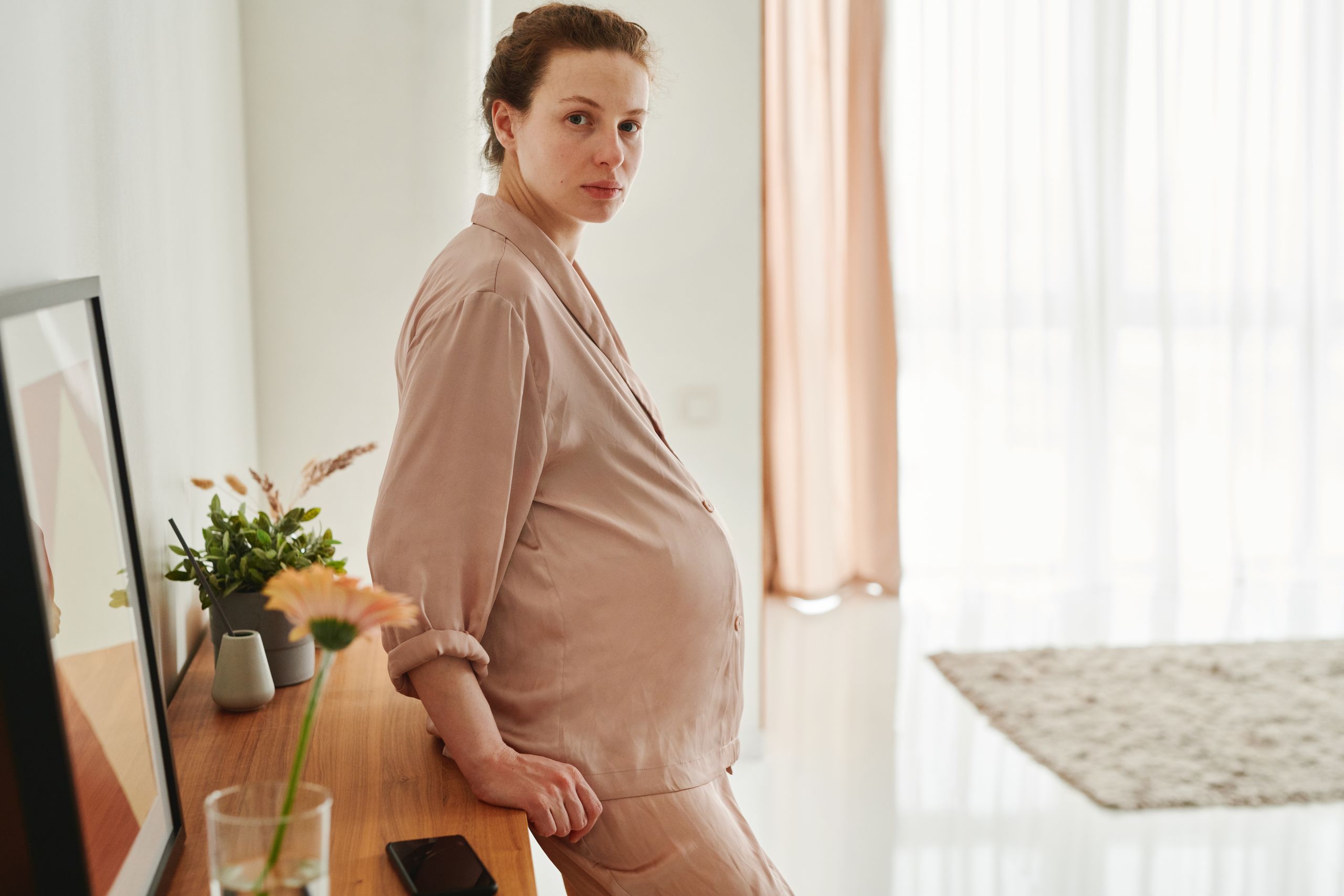PGD and IVF 6 A pregnant woman wearing sleepwear leans on a wooden drawer in a bright, serene room.